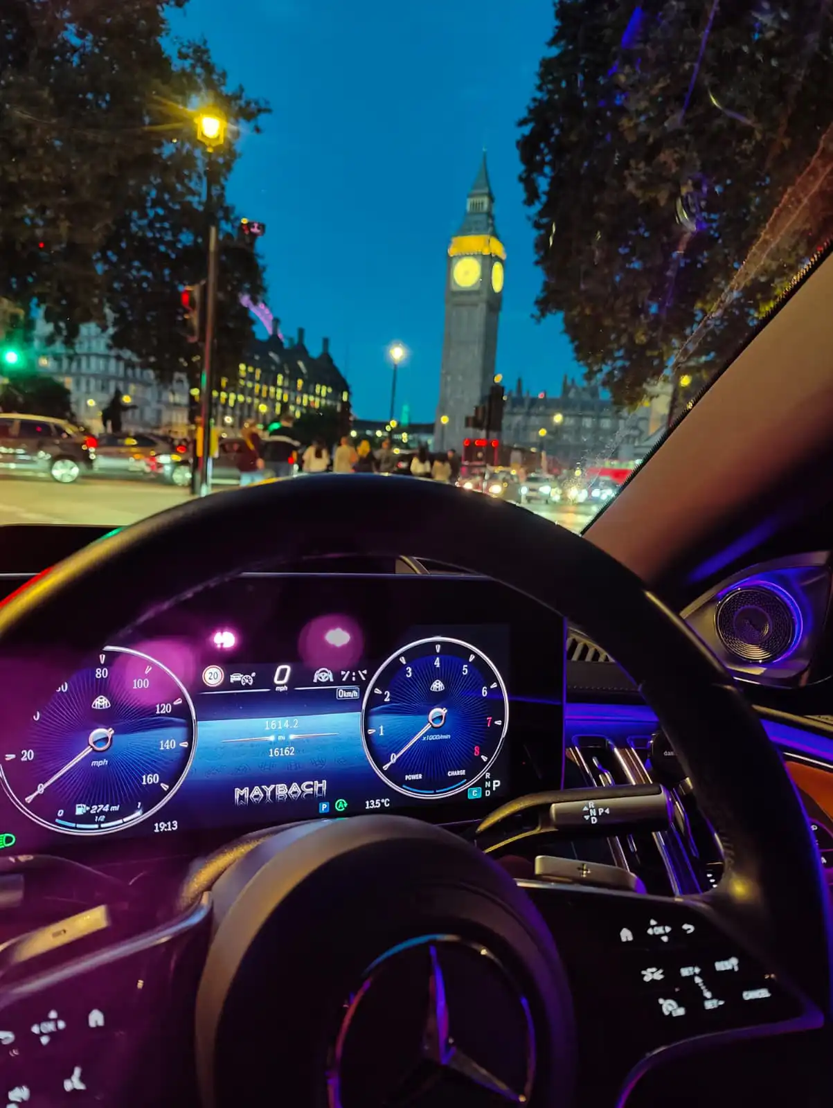 Interior view of a luxury Mercedes-Maybach chauffeur car dashboard with ambient lighting, looking through the windshield at the illuminated Big Ben clock tower at dusk in London.