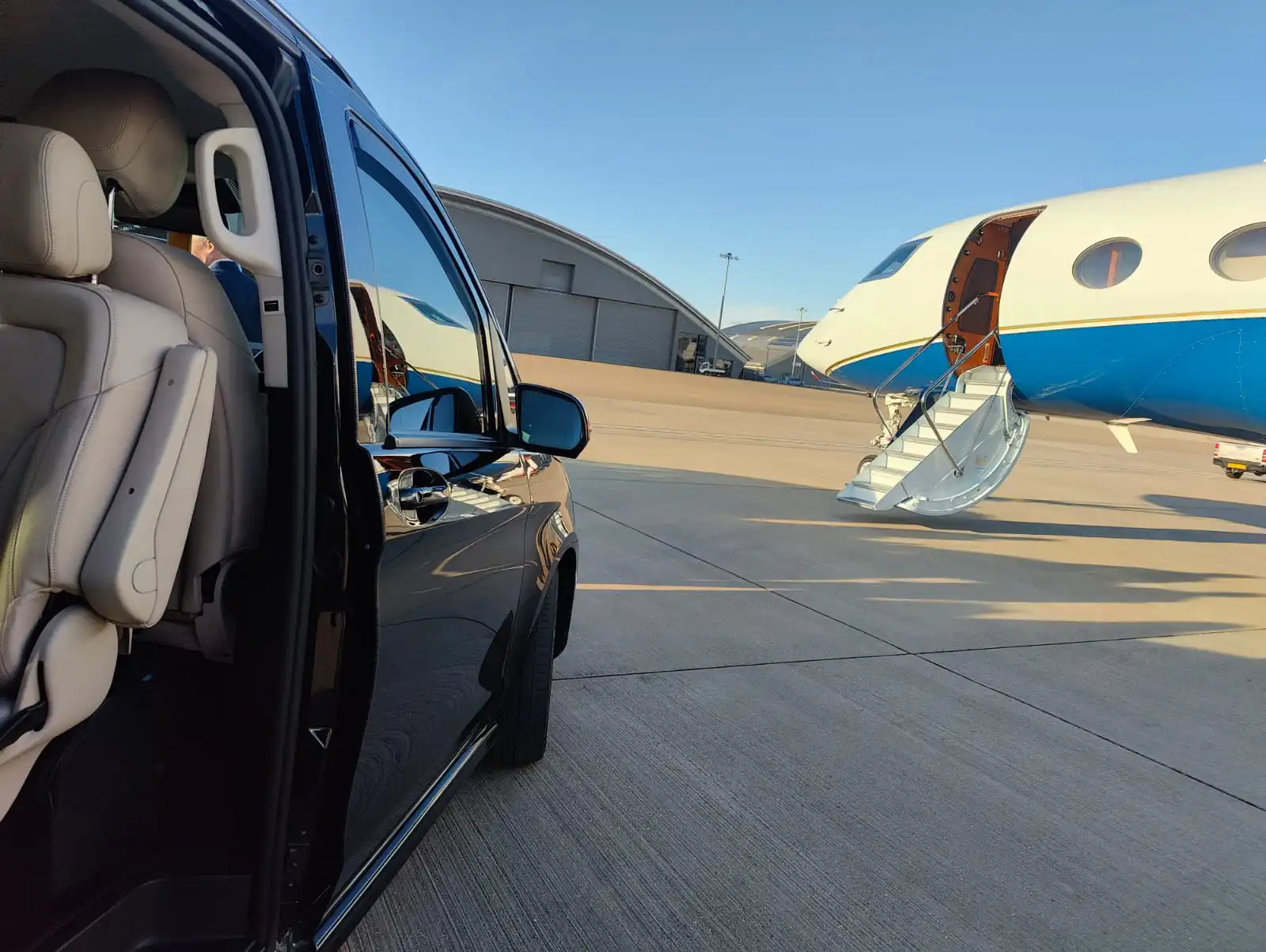 A black Mercedes-Benz V-Class chauffeur MPV with its passenger door open, parked on the airport tarmac directly next to the steps of a private jet.