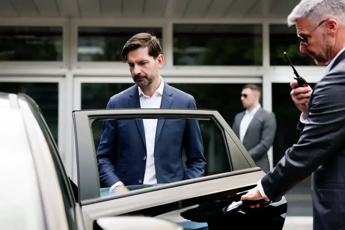 A security chauffeur holds a black car door open for a man in a blue suit. The chauffeur speaks into a radio. A second security guard stands in the background.