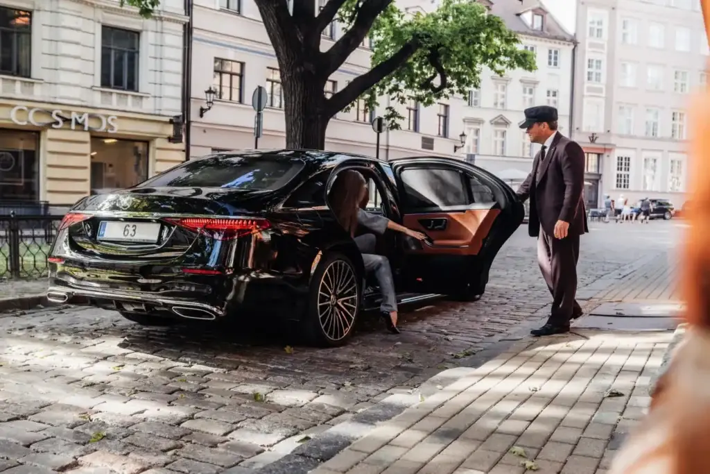 A professional chauffeur wearing a dark suit and a traditional cap holds open the rear door of a black Mercedes-Benz S-Class on a cobbled city street as a female passenger steps inside the luxury tan leather cabin.