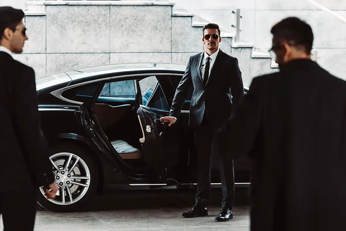 A security chauffeur in a black suit and sunglasses holds open the rear door of a luxury black saloon car. Two additional close protection officers stand guard in the foreground.